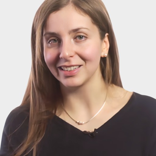 A woman with long brown hair wearing a black top and a necklace, smiling at the camera against a plain light background.