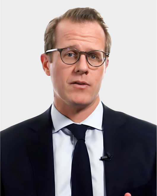 A man wearing glasses, a dark suit, white shirt, and navy tie stands against a plain light background, looking directly at the camera.