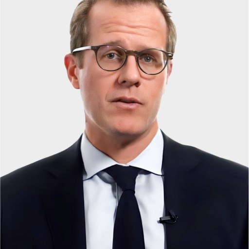 A man wearing glasses, a dark suit, white shirt, and navy tie stands against a plain light background, looking directly at the camera.