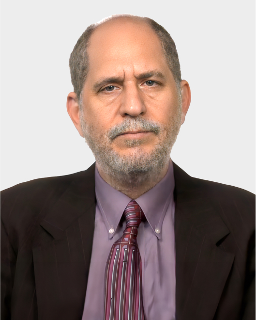 A middle-aged man with short gray hair and a beard, wearing a dark suit, lavender shirt, and striped tie, poses against a plain light background.