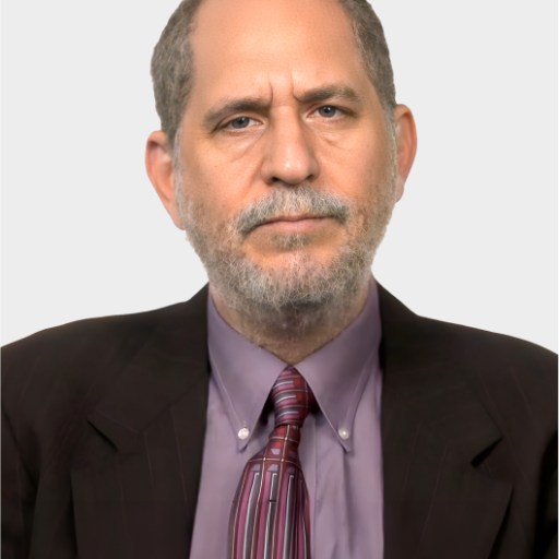 A middle-aged man with short gray hair and a beard, wearing a dark suit, lavender shirt, and striped tie, poses against a plain light background.